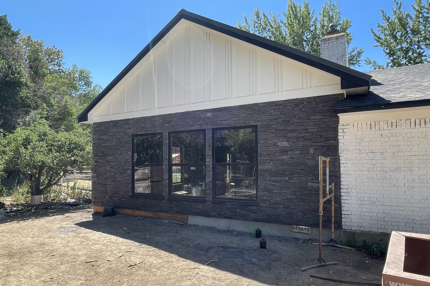 Home addition under construction with dark stacked stone, black-framed windows, and a white board and batten gable